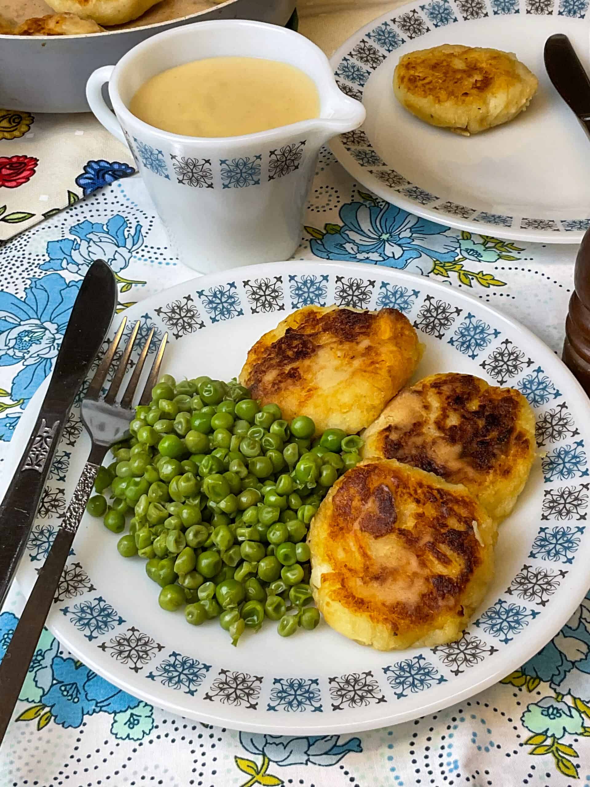 Parsnip patties served on a vintage patterned plate with peas and fork and knife to side, small jug of cheese sauce in background.