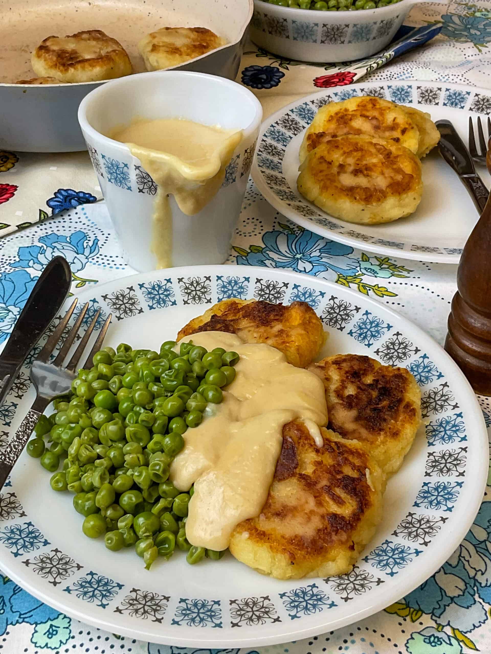 Parsnip patties served with peas and cheese sauce, cheese sauce jug to side with a cheese drip down the side.