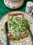 Cheesy peas on toast served on lemon patterned plate with fork and knife to side, flower patterned pot in background and a green table cloth.